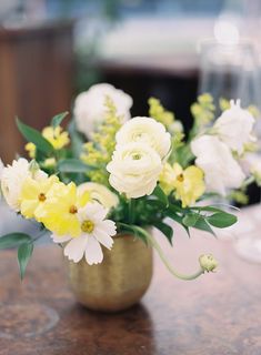 a vase filled with white and yellow flowers on top of a wooden table next to wine glasses