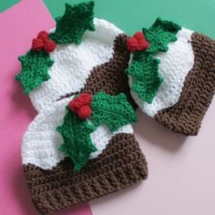 two crocheted hats with holly leaves on them sitting on a pink and green surface