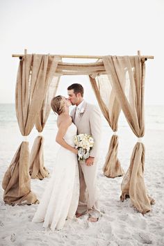 a bride and groom kissing on the beach in front of an outdoor wedding ceremony arch