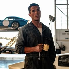 a man in overalls holding a coffee cup and looking at the camera while standing next to a car