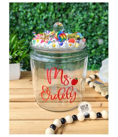 a glass jar filled with candy on top of a wooden table next to a green bush