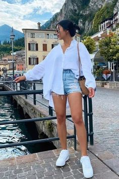 a woman standing next to a body of water wearing white sneakers and a white shirt