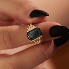 a woman's hand with black nail polish holding a gold and green stone ring