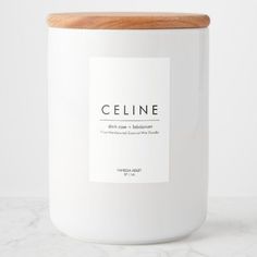 a white candle with a wooden lid on a marble countertop next to a bottle of celine