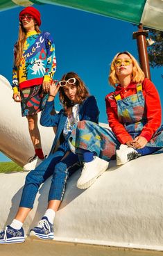 three young women sitting on top of a white structure wearing colorful clothing and sunglasses, all looking at the camera