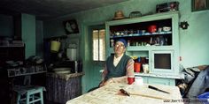 an old woman sitting at a table in a room with many things on the shelves