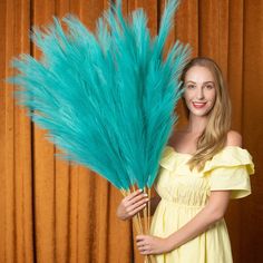 a woman in a yellow dress is holding a large blue feather plant and smiling at the camera