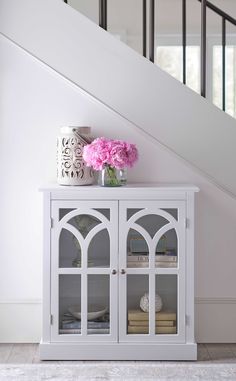 a white cabinet with glass doors and pink flowers on top in front of a stair case