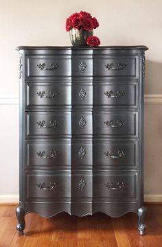 a silver dresser with flowers on top of it in front of a white wall and wooden floor