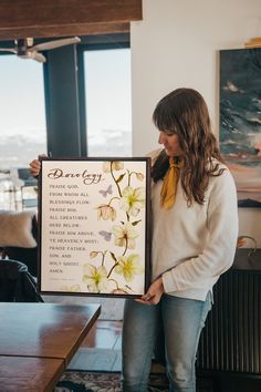 a woman holding up a framed sign with flowers on it in front of a window