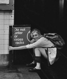 a black and white photo of a woman leaning against a sign that says do not enter or cross tracks