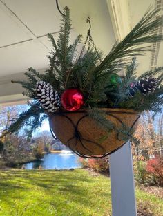 a hanging basket filled with pine cones and christmas decorations on a porch next to a lake