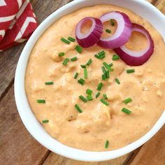 a white bowl filled with dip topped with onion slices and sliced onions next to a red napkin