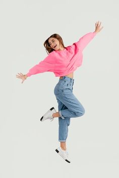 a woman jumping in the air with her arms out and legs spread wide, wearing jeans and a pink sweater