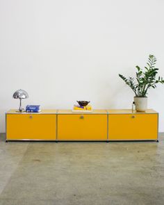 a yellow sideboard with two plants on top and a lamp next to it in front of a white wall