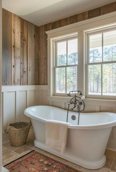 a white bath tub sitting under two windows in a bathroom next to a rug on the floor