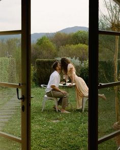 a man and woman sitting at a table in front of an open window, kissing