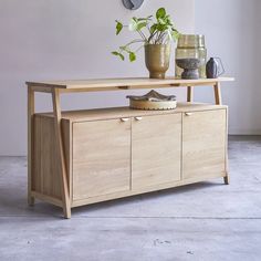 a wooden cabinet with two plants on top and a clock hanging above it, next to a potted plant