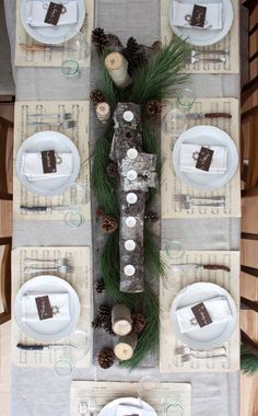 the table is set with white plates and silverware, pine cones, and candles