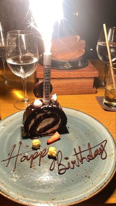 a piece of cake on a plate with the word happy birthday written on it and some wine glasses in the background