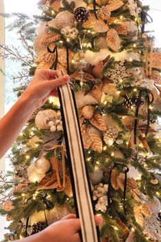 a christmas tree decorated with gold and white ornaments, ribbons and pine cones is being held up by someone's hand