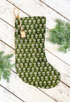 a green stocking with white flowers on it sitting next to evergreen branches and pine cones