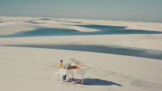 two people sitting at a white piano in the middle of an expanse of sand and water