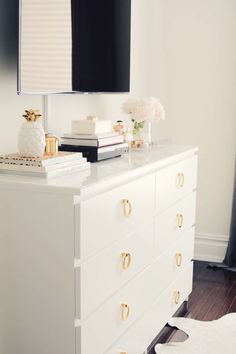 a white dresser with gold handles and some books on top of it in front of a flat screen tv