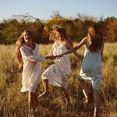 three girls are holding hands and running through tall grass in a field with trees in the background