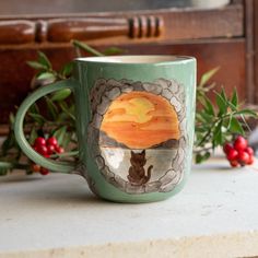 a green coffee mug with a cat sitting on top of it next to holly branches