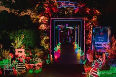 an entrance to a garden at night with neon lights on the walkway and potted plants