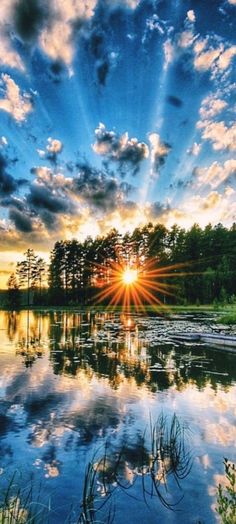 the sun is setting over a body of water with trees in the background and clouds in the sky