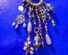 Art Nouveau inspired plum, lavender, and deep purple gems create a fresh lush pendant.  Cascading from a bold one inch brass ring are multi shapes and sizes of amethyst, agate, clear crystal quartz, hematite, with floral and heart charms.  This 4 inch long Boho style pendant is Artisan Handcrafted in America and has a 36 inch black silk cord that can easily be made shorter.  If you prefer, I am happy to custom make a chain with gemstones, or other colored silk or leather cord.  Enjoy! Inv #  P425 Retail $98 SHOP POLICIES: QUALITY & VALUE: Fresh Jewels are Artisan Handcrafted in America, designed to enjoy wearing for a lifetime, and may become a cherished heirloom. Prices here are up to 75% OFF prices compared to Designer Stores and Boutiques! Every Jewel is backed with a Quality Guarantee, Purple Pendant Necklaces With Charms, Purple Pendant Necklace With Charms, Handmade Artistic Purple Necklace, Artistic Handmade Purple Necklaces, Unique Purple Dangle Necklaces, Purple Bohemian Jewelry With Lobster Clasp, Purple Jewelry With Dangling Beads For Gift, Gift Purple Jewelry With Dangling Beads, Lavender Dangling Beads For Jewelry Making