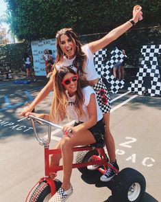 two girls are riding on a tricycle in the street with their arms out and one girl is wearing red sunglasses