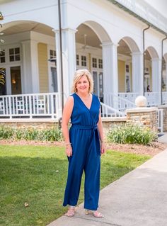 a woman standing in front of a white house wearing a blue jumpsuit and sandals