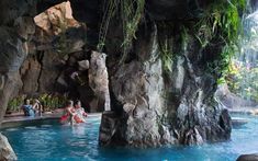 some people are sitting in the water next to large rocks and plants, while others look on