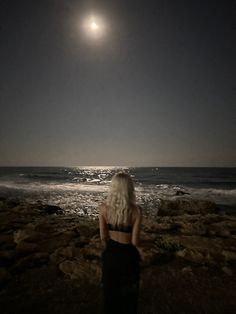 a woman standing on top of a rocky beach next to the ocean under a full moon