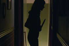 the silhouette of a woman holding a knife is seen through an open door at night