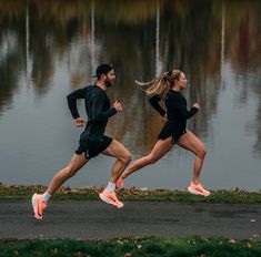 two people are running in the street near some water with trees and grass behind them