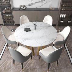 a white marble dining table surrounded by grey chairs and a black book shelf with wine bottles on it