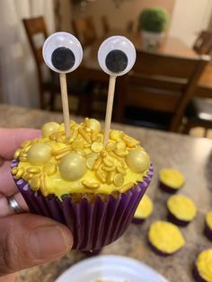 a cupcake with googly eyes and yellow icing is held up in front of the camera