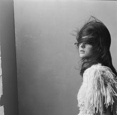 black and white photograph of a woman with long hair wearing a feathered coat, standing in front of a wall