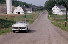 an old car is driving down the road in front of a farm house and silo