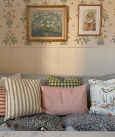 a couch with several pillows on it in front of a wallpapered wall and framed pictures