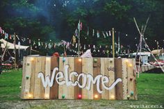 a welcome sign with lights on it in front of some tents and flags hanging from the trees