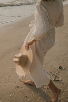 a woman is walking on the beach with a hat in her hand and wearing a white dress