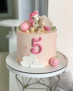 a pink birthday cake with a unicorn on top and clouds around it, sitting on a table