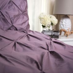 a bed with purple sheets and pillows in a room next to a lamp on a table