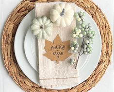 a place setting on a wicker plate with white pumpkins and greenery in the center