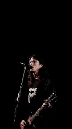 a man with long hair holding a guitar and singing into a microphone while standing in front of a black background